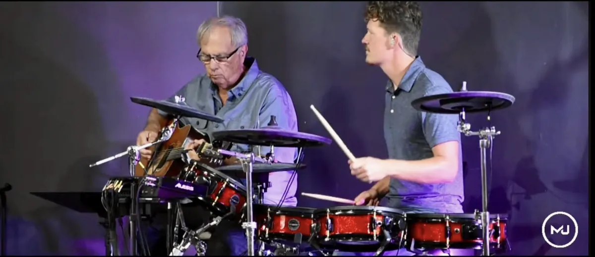Two drummers, one older and one younger, playing side by side.