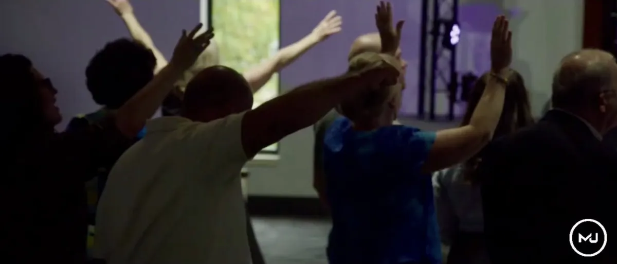 Worshippers seen from behind with their arms raised in violet stage light.