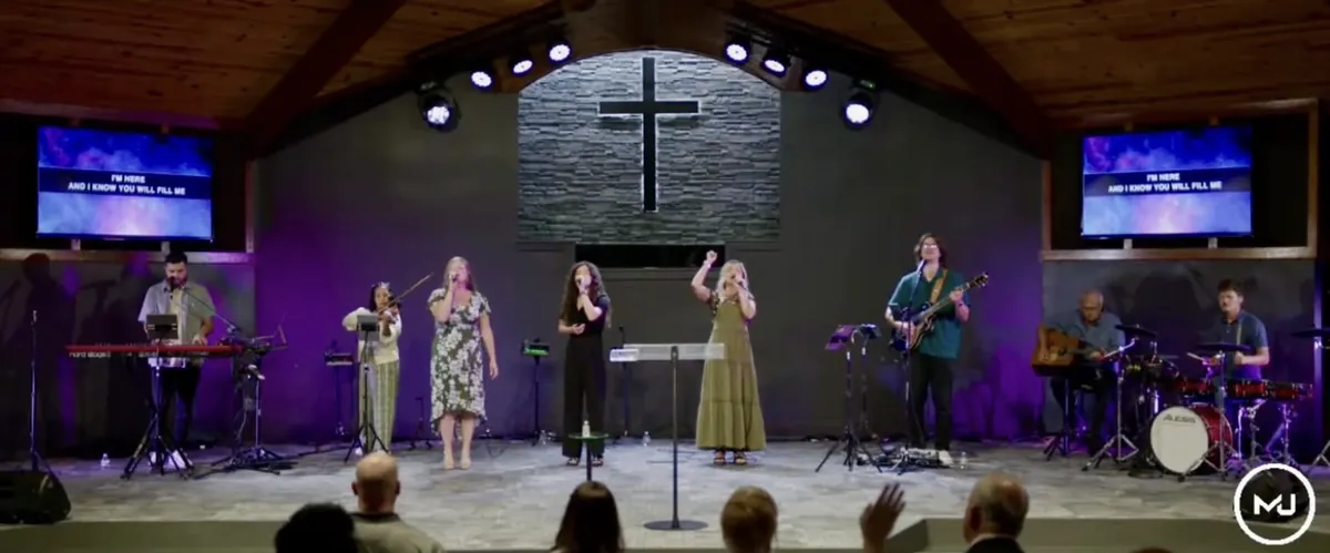 The worship band on stage beneath a backlit cross on a stacked-stone wall, with lyric screens on both sides of the sanctuary